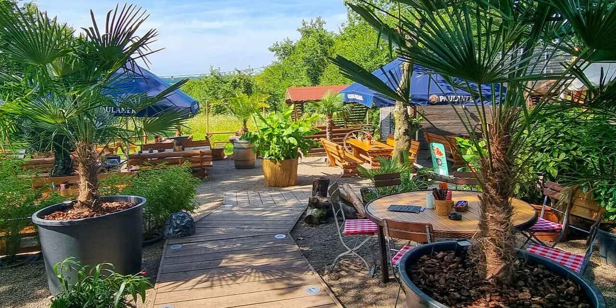Holzsteg durch den Biergarten, gesäumt von Palmen und Kübelpflanzen