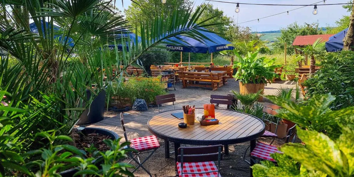 Runder Tisch im Biergarten unter Lichterketten, ringsum Palmen und Grünpflanzen