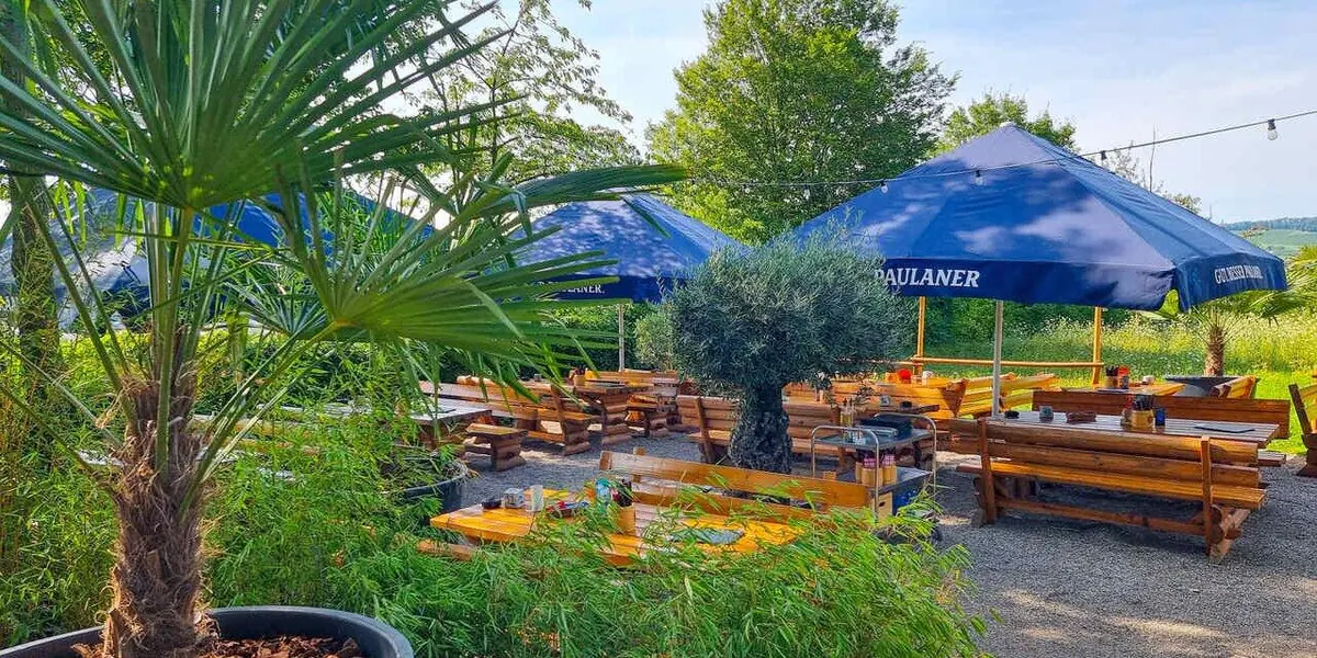 Biergarten mit blauen Sonnenschirmen und langen Holzbänken zwischen Palmen